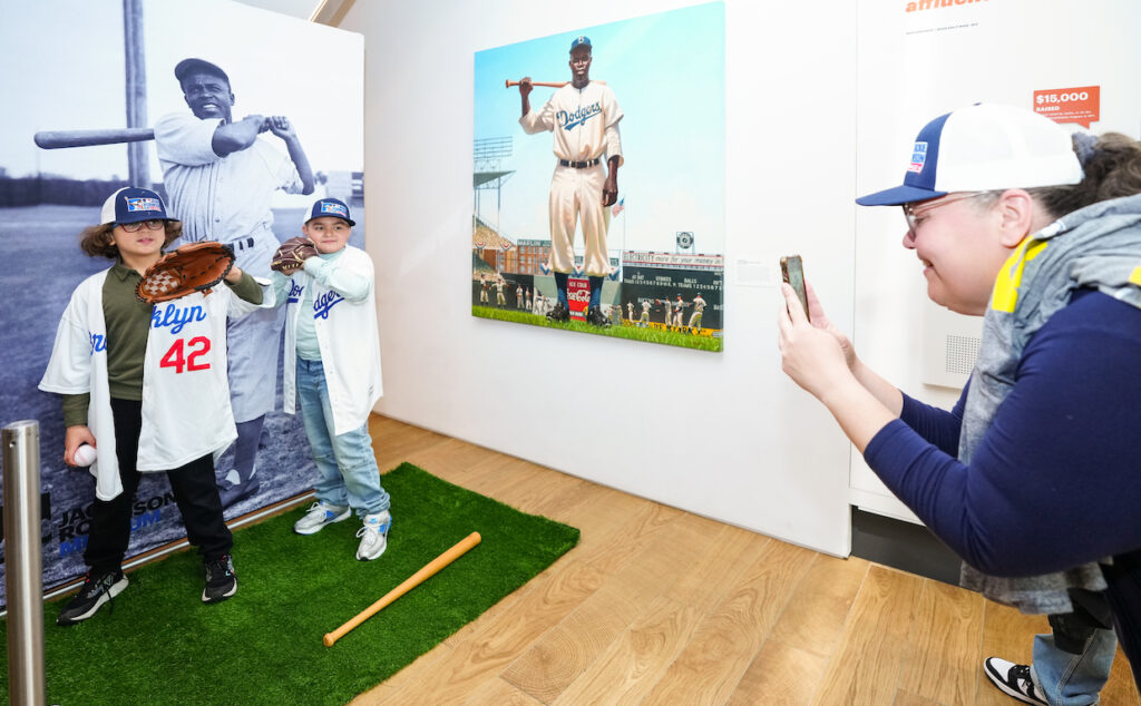 Two children pose with baseball props in front of a life size photo of Jackie Robinson while an adult takes a photo.