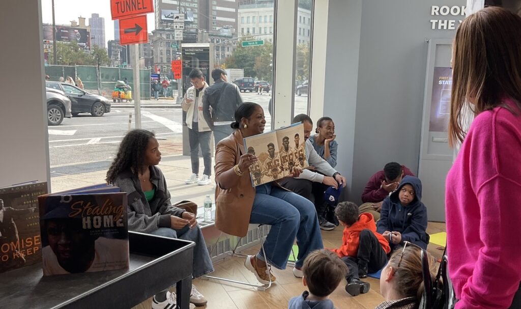Sonya Pankey Robinson is seated holding a picture book open to an illustration of Negro League baseball players surrounded by children and adults seated on the floor.
