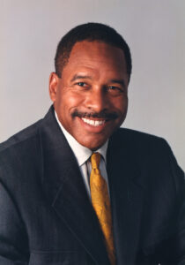 Headshot against grey background wearing dark suit and yellow tie.
