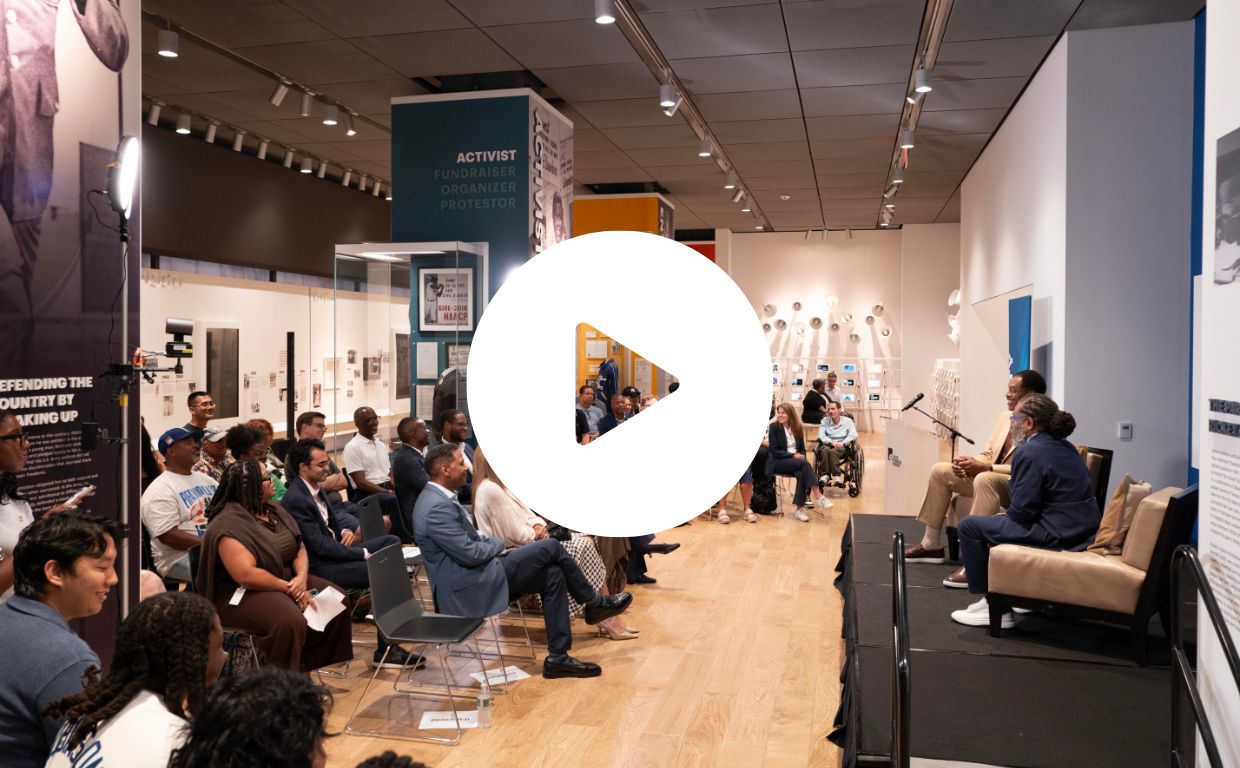A view from stage left of two speakers on a stage at the right and the audience seated in a museum gallery on the left.